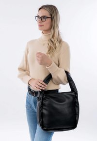 Donna con capelli biondi e occhiali, che indossa un maglione beige e jeans, mentre tiene una grande borsa in pelle nera con manici imbottiti.