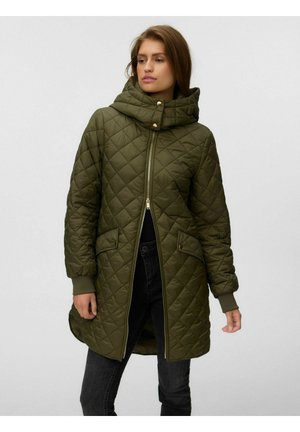 Femme portant une veste matelassée vert olive avec col haut, fermeture éclair sur le devant, poches latérales à boutons et poignets côtelés, debout devant un fond uni.