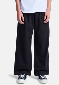 Schwarze weite Hosen aus Baumwolle mit lockerem Schnitt und umgeschlagenen Säumen, kombiniert mit schwarz-weißen Segeltuchschuhen.