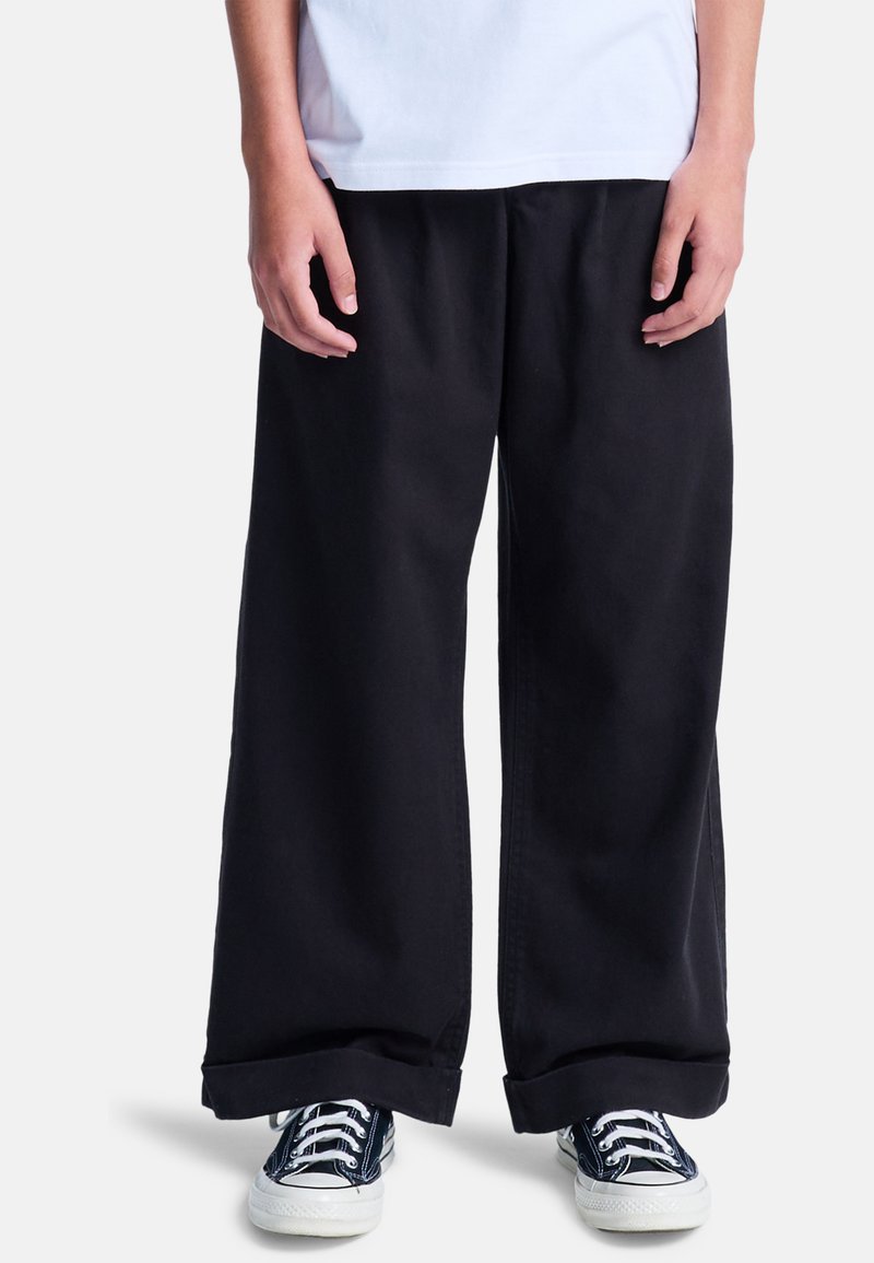 Schwarze weite Hosen aus Baumwolle mit lockerem Schnitt und umgeschlagenen Säumen, kombiniert mit schwarz-weißen Segeltuchschuhen.
