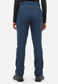 Osoba ubrana w granatowe spodnie turystyczne i czarne buty trekkingowe, stojąca tyłem do przodu na jednolitym tle.