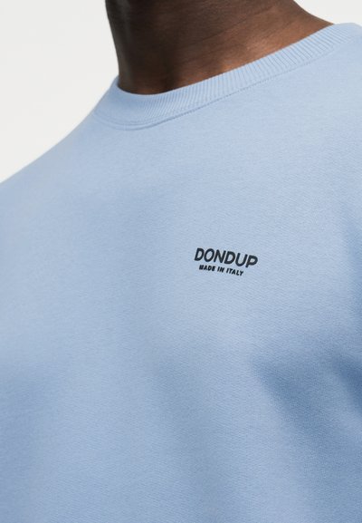 Κοντινό πλάνο ατόμου που φοράει ανοιχτό γαλάζιο φούτερ με μαύρο κείμενο "DONDUP MADE IN ITALY" στο στήθος.