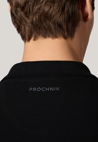Czarna koszulka polo z teksturowanej tkaniny, z szarym logo z napisem "PROCHNIK" na tylnej części kołnierza. Prosty design z klasycznym krojem.
