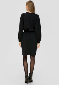 Czarna sukienka z długimi rękawami, dopasowana w talii, o długości do kolan, wykonana z miękkiego materiału. Noszona z czarnymi botkami za kostkę i przezroczystymi rajstopami.
