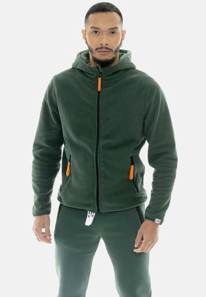 Homme portant une veste polaire vert foncé avec des tirettes de fermeture éclair orange et un pantalon vert assorti, debout devant un fond uni.