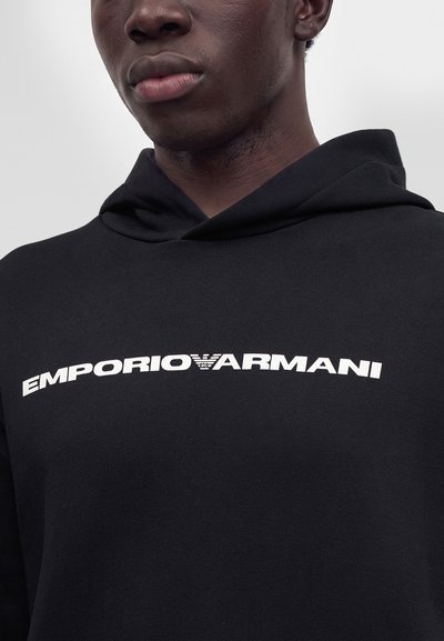Крупен план на човек, носещ черна блуза с качулка с бяло лого Emporio Armani, центрирано на гърдите.