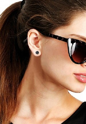Lunettes de soleil avec une monture écaille de tortue, verres teintés foncés, portées avec un haut noir. Boucles d'oreilles à clous noires visibles à l'oreille. Cheveux lisses en queue de cheval.