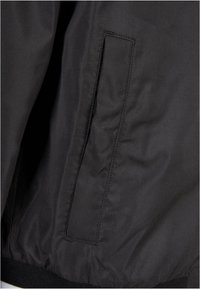 Chaqueta negra con textura suave, puños elásticos y un bolsillo lateral. El tejido parece ligero y resistente al agua.