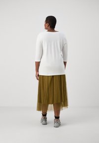 Top branco de manga três quartos combinado com uma saia pregueada verde-oliva. O modelo usa ténis cinzentos com detalhes em preto.