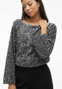 Grijze pailletten top met een losse pasvorm, ronde halslijn en lange mouwen. De oppervlaktextuur is glanzend en reflecterend, wat visuele interesse toevoegt.