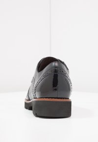 Scarpa in pelle verniciata nera con dettagli brogue, punta arrotondata e tacco in legno impilato. Texture liscia con perforazioni decorative.
