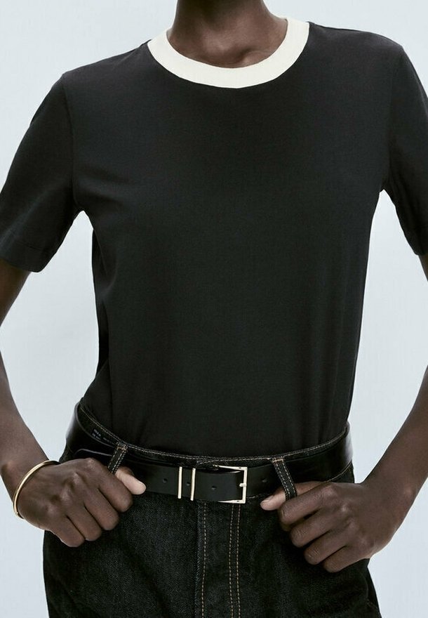 Une personne porte un t-shirt noir avec un col blanc et un jean noir, tenant la boucle de ceinture en cuir noir avec les deux mains.