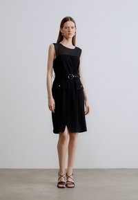 Sort kjole med et gjennomsiktig øverste lag, til knelengde, med belte i midjen og sidelommer. Brukes med svarte remmesandaletter.