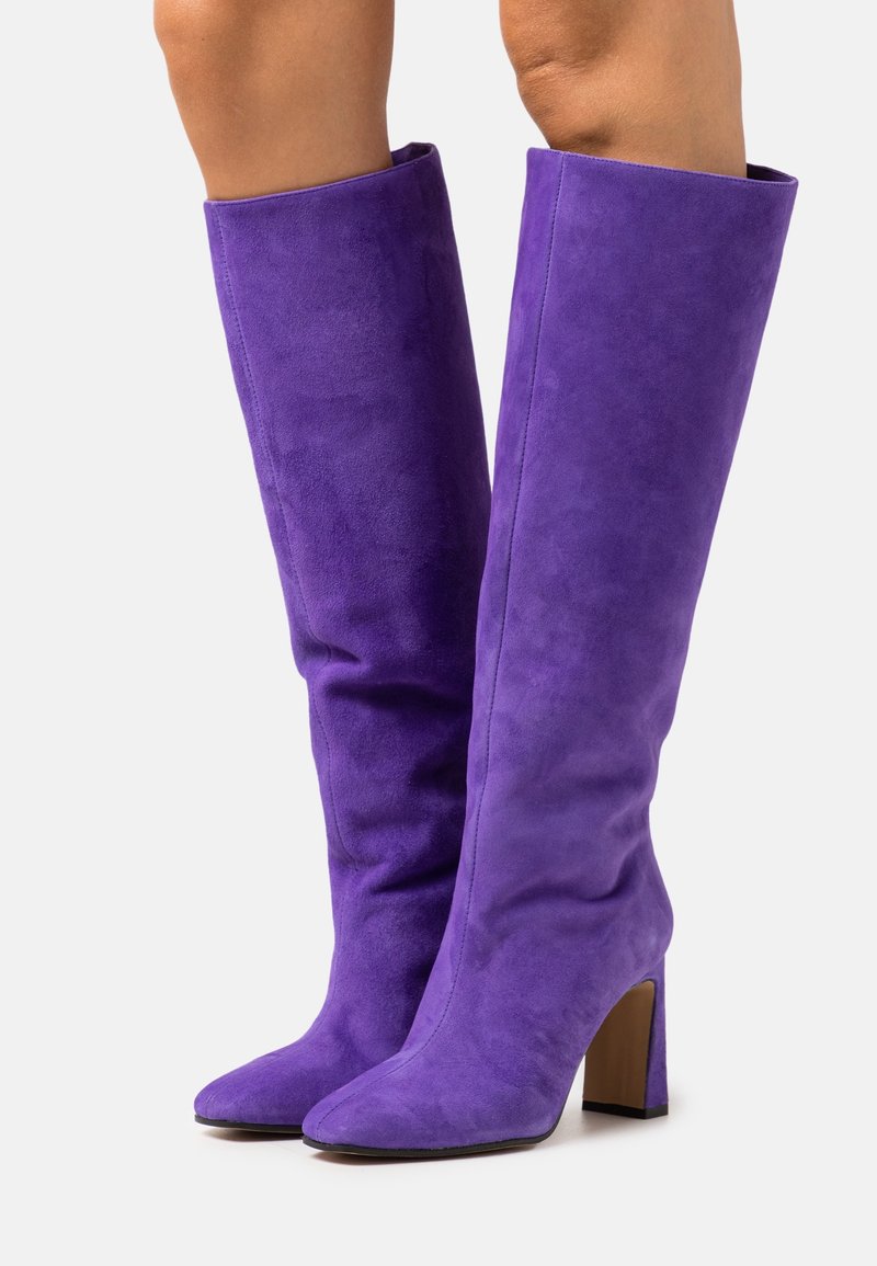 Bottes hautes en daim violet à bouts pointus et talons blocs portées sur des jambes nues sur fond blanc.
