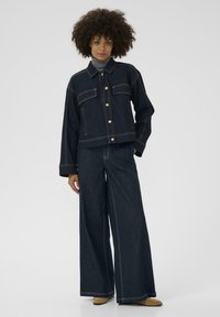 Tumman denim-kankaan takki, jossa on kultaiset napit ja kaksi etutaskua, yhdistettynä leveälahkeisiin tumman denim-kankaan housuihin. Sujuva tekstuuri ja hyvin istuva malli.