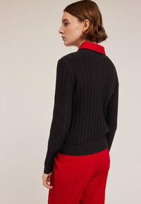 Czarny żebrowany sweter z długimi rękawami i dopasowanym krojem, z czerwonym kołnierzykiem. Noszony z czerwonymi spodniami. Teksturowana tkanina.