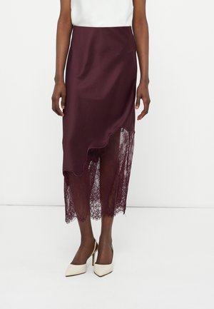 Vrouw draagt een bordeauxrode midi-rok met asymmetrische, geschulpte zoom en een kanten overlay, gecombineerd met witte pumps met spitse neus.