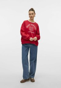 Sweatshirt rouge avec "SAN DIEGO 86 CALIFORNIA" imprimé en blanc. Assorti avec un jean bleu ample et des baskets marron. Texture lisse, coupe décontractée.