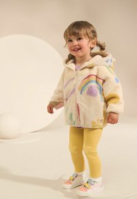 Veste en polaire avec un design de arc-en-ciel coloré, couleur crème, fermeture éclair, associée à des leggings jaunes avec des accents d'étoiles et des baskets multicolores.