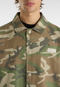 Tarnjacke mit einer Knopfleiste vorne, aus robustem Stoff. Verfügt über Farbtöne in Grün, Braun und Beige, mit einem kleinen "VANS"-Logo-Patch.