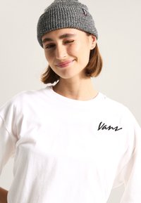 Gorro de punto gris, textura acanalada, con una pequeña etiqueta de logo. Camiseta blanca con el logo negro de "Vans". Diseño simple y casual, ajuste relajado.