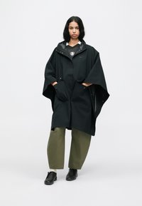 Naine seisab seljas must suur kapuutsiga ponšo, rohelised lai jalad püksid ja mustad kingad, üksluisel valgel taustal.