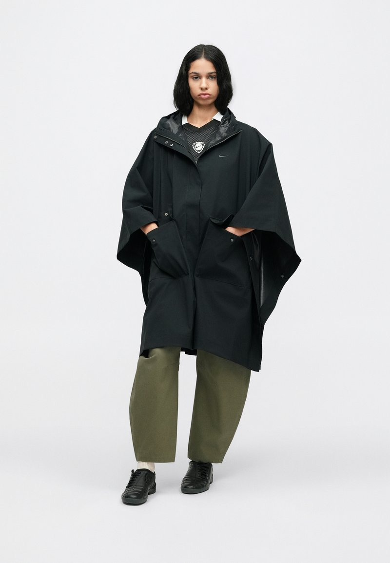 Woman standing wearing a black oversized hooded poncho, green wide-leg pants, and black shoes against a plain white background.