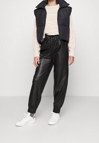 Black padded vest, cream cable-knit sweater, black faux leather joggers with elastic cuffs, and white sneakers. Casual outfit with mixed textures.