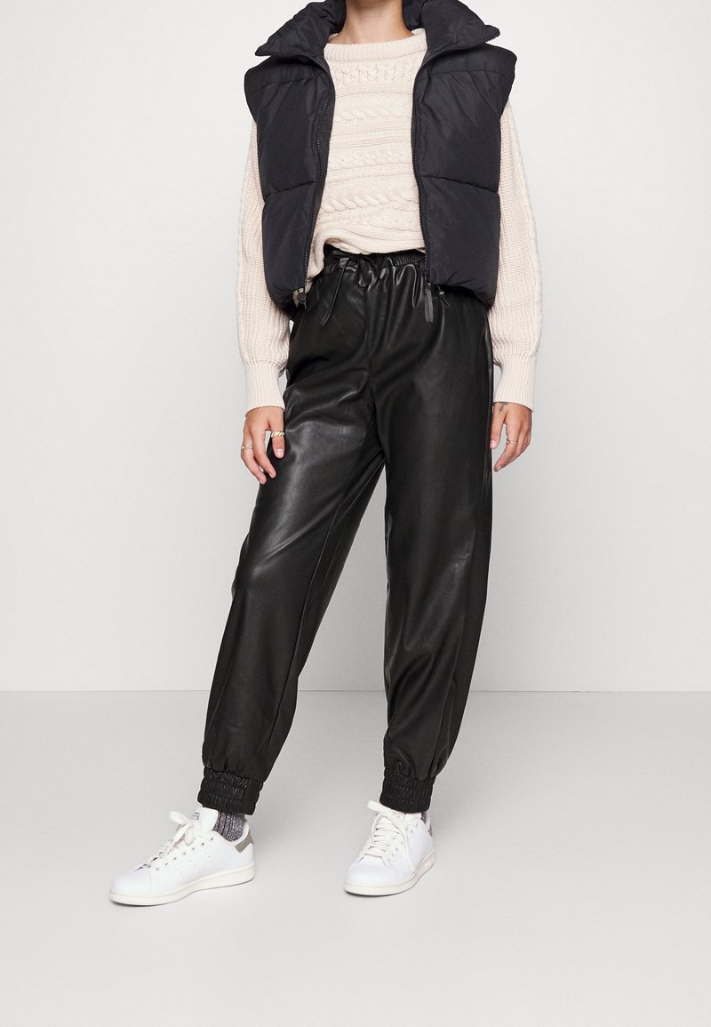 Black padded vest, cream cable-knit sweater, black faux leather joggers with elastic cuffs, and white sneakers. Casual outfit with mixed textures.