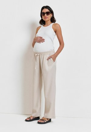 Zwangere vrouw draagt een wit tanktopje, beige losse broek, zwarte sandalen en een zonnebril, staat met één hand op haar buik en één in haar broekzak.
