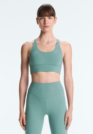 Femme portant un soutien-gorge de sport vert sauge et des leggings taille haute, debout droite contre un fond clair uni.