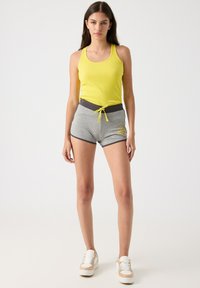 Top sin mangas amarillo acanalado con tirantes delgados, combinado con pantalones cortos grises que tienen un cordón y un logo amarillo. Zapatillas blancas adjuntas completan el look.
