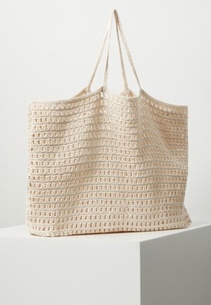 Sac cabas beige tricoté avec motif en maille ouverte et deux fines bretelles reposant sur un piédestal blanc sur un fond uni.
