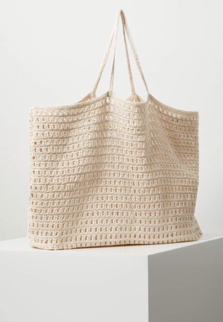 Sac cabas beige tricoté avec motif en maille ouverte et deux fines bretelles reposant sur un piédestal blanc sur un fond uni.