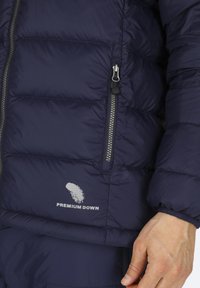 Marinblå pufferjacka med en dragkedjeficka, kviltat mönster och en "Premium Down"-logotyp tryckt i vitt. Elastiska muddarna är synliga.