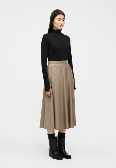 Γυ ναίκα που στέκεται φορώντας μαύρο μακρυμάνικο τοπ, μπεζ μίντι φούστα, μαύρες κάλτσες και μαύρα δερμάτινα μποτάκια στον αστράγαλο, σε απλό λευκό φόντο.