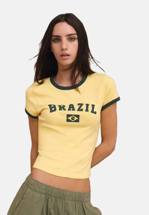 Giovane donna con capelli lunghi e scuri che indossa una maglietta gialla corta con rifiniture verdi e la scritta "BRAZIL" con la bandiera, abbinata a pantaloni verde oliva.