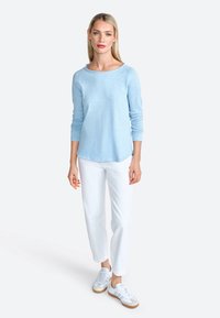Camicia azzurra a maniche lunghe con scollatura rotonda, abbinata a pantaloni bianchi e sneakers bianchi con suole marroni. Texture liscia.