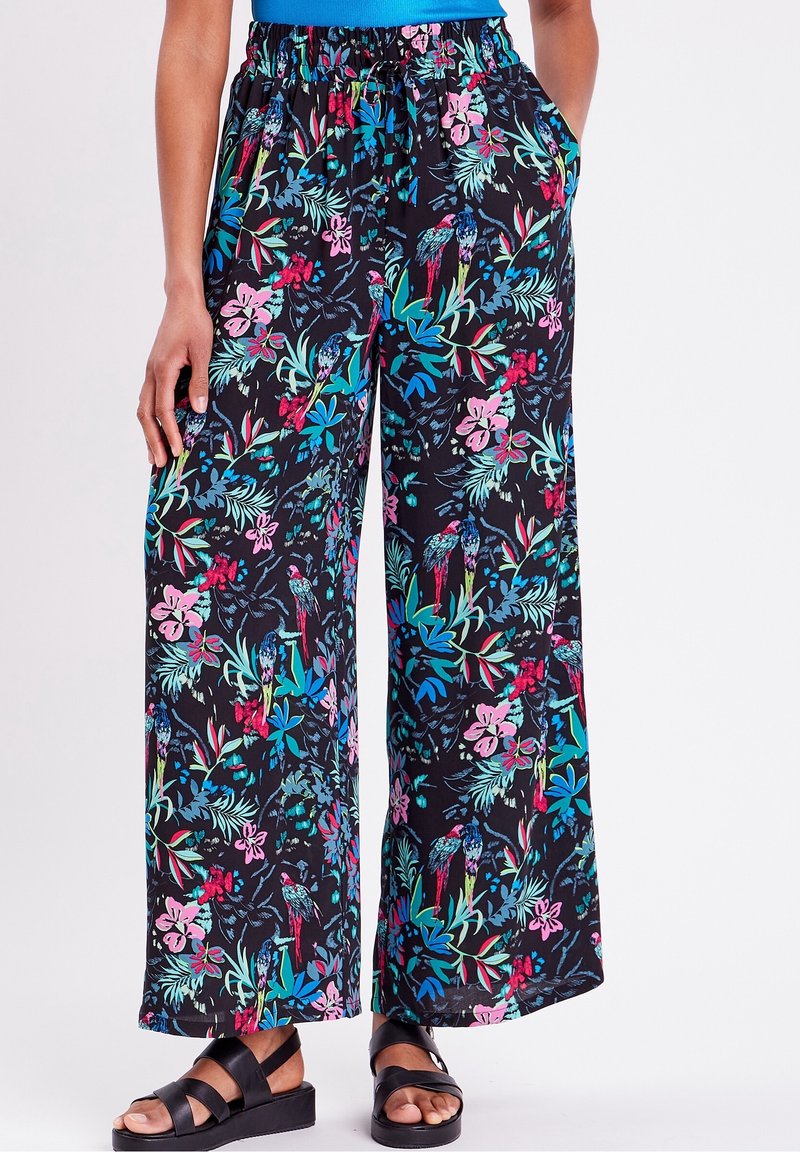 Pantalon noir à jambes larges avec imprimé tropical coloré de fleurs et d'oiseaux, taille élastique, porté avec des sandales noires et un haut bleu.