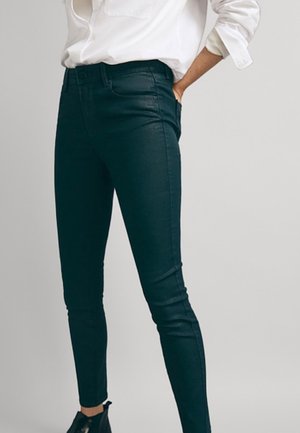 Femme portant un jean slim noir, une chemise blanche à manches longues avec les manches retroussées, et des bottines noires sur un fond uni.