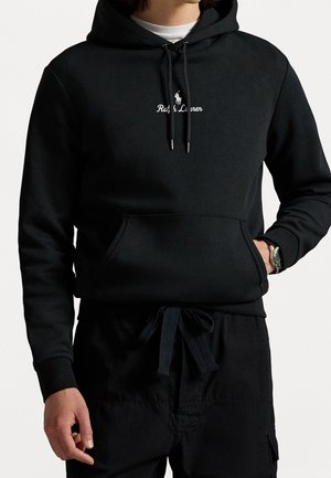 Mand iført sort Ralph Lauren hættetrøje med forlomme, snore og sorte bukser, hånd i lommen, hvid skjorte synlig under hættetrøjen.