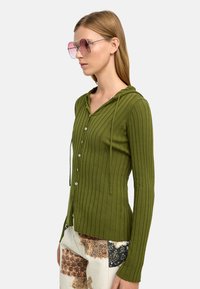 Zielony kardigan w prążki z kapturem, z przednim zapięciem na guziki i długimi rękawami. Noszony z wzorzystymi, jasnymi spodniami i dużymi różowymi okularami przeciwsłonecznymi.