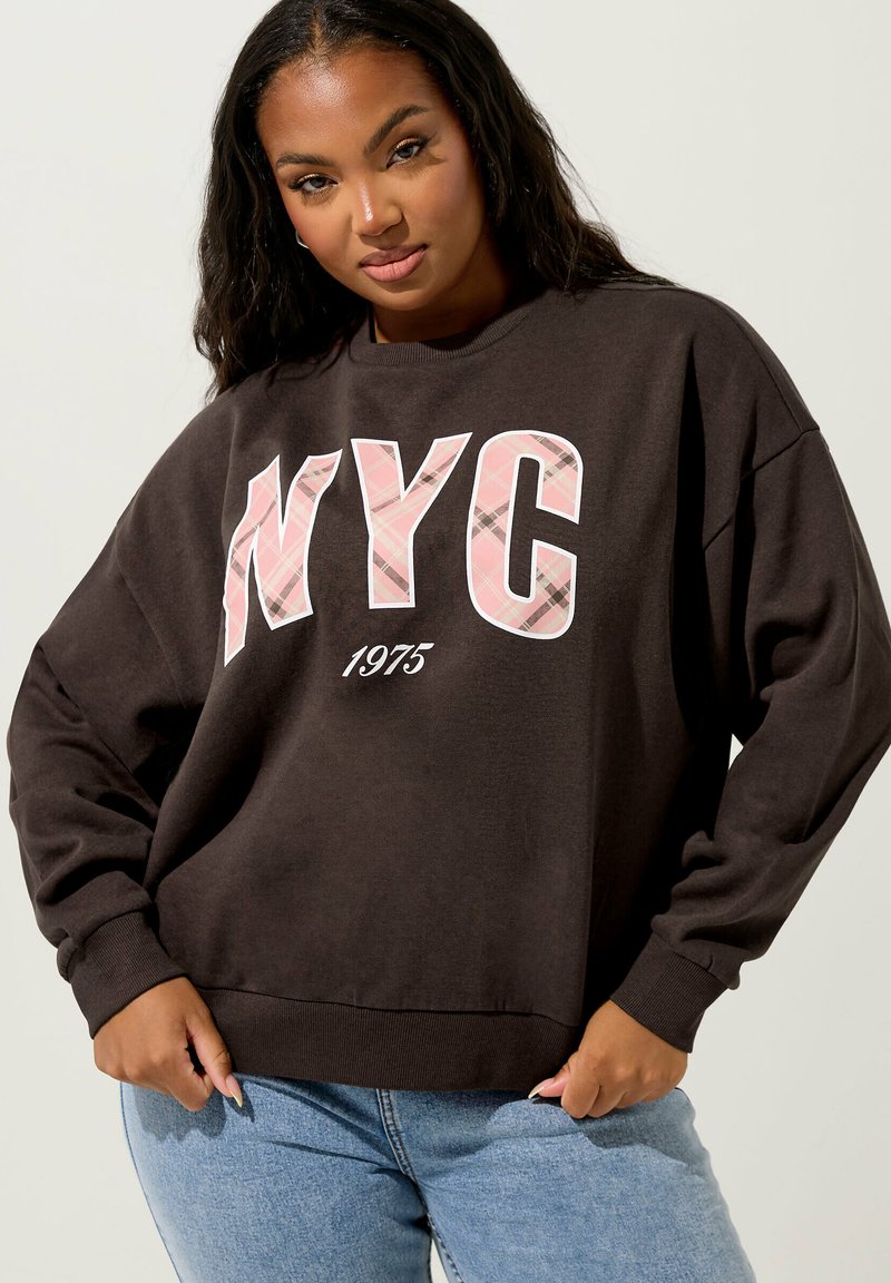 Felpa oversized marrone con grafica a quadri bianchi e rosa "NYC" e testo "1975". Polsini e orlo a coste, vestibilità ampia, tessuto morbido.