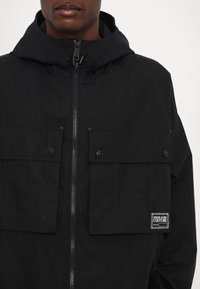 Black jacket with a zip front, large flap pockets, adjustable hood, and textured fabric. Features a logo patch on the left pocket.