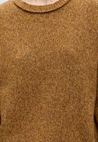 Gros plan d'une personne portant un pull en maille marron texturé à col rond, montrant uniquement le haut de la poitrine et les épaules.