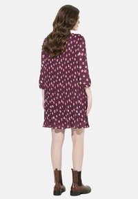 Lila gefaltetes Kleid mit kleinen weißen und rosa Blumenmustern, 3/4-Ärmel und lässiger Passform, kombiniert mit braunen Stiefeletten.