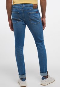 Siniset farkut, joissa on slim fit -leikkaus, kaksi takataskua, vaaleat tikkausompeleet ja käännetty lahje. Yhdistetty valkoisiin ja sinisiin lenkkareihin.