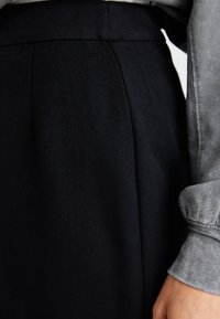 Saia de tecido preto com uma textura suave, apresentando uma cintura limpa e pregas frontais discretas. Camisa cinza com punhos texturizados visíveis.