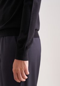 Czarny sweter dzianinowy z dopasowaną talią i długimi, teksturowanymi rękawami, zestawiony z ciemnymi spodniami. Zbliżenie na ramię i dłoń.