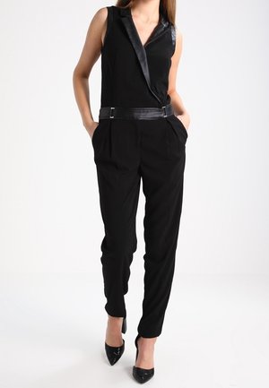 Vrouw in een mouwloze zwarte jumpsuit met leren revers en riem, gecombineerd met zwarte puntige hoge hakken, staande met handen in de zakken.